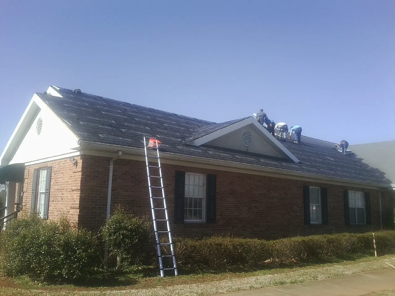 Crew tearing shingles off a brick ranch roof with a ladder set — Metal Roof Repair work by our crew in Schwertner