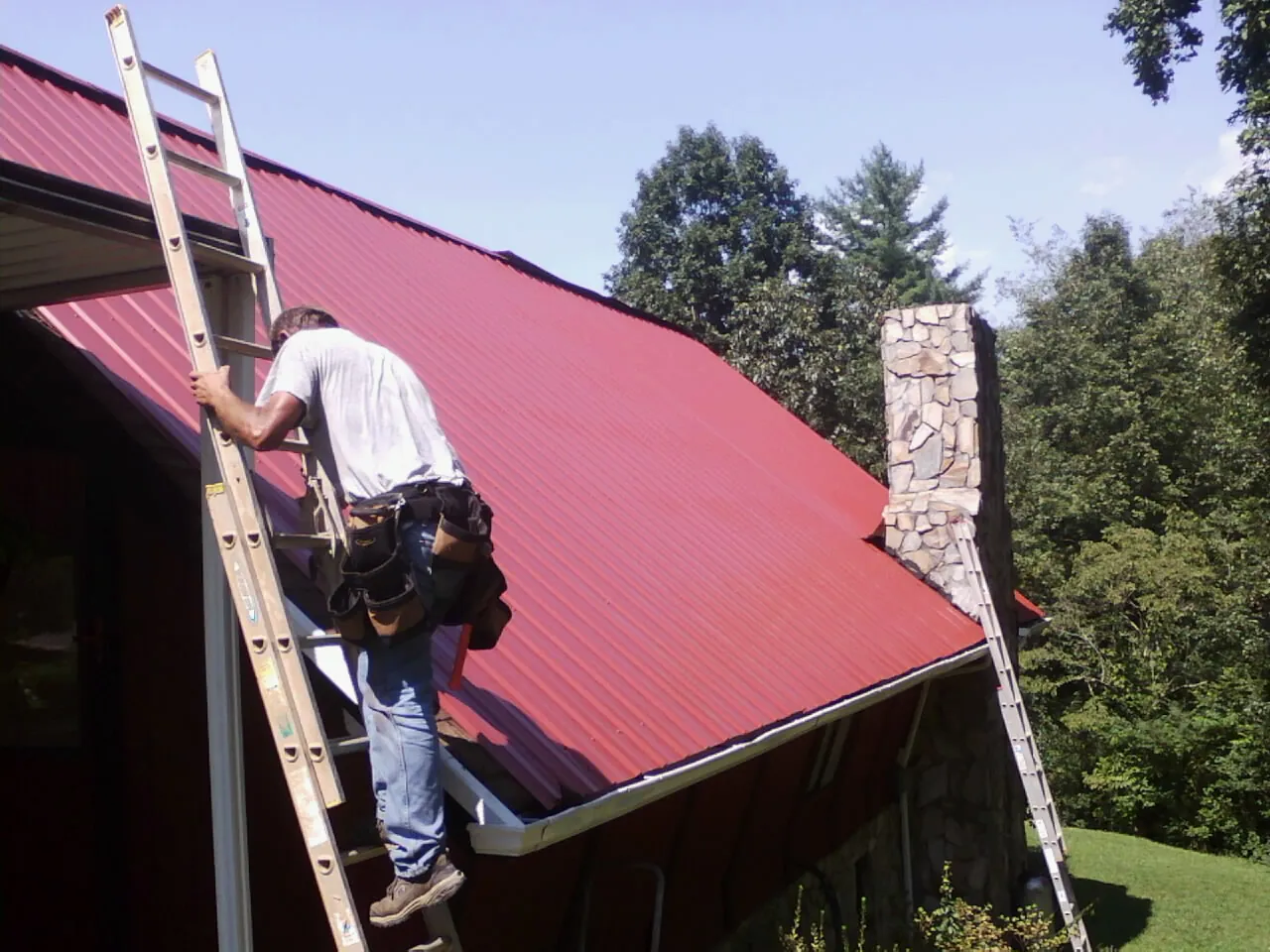 Roofer on a ladder beside a red metal roof and stone chimney — Soffit & Fascia Repair by Roofing Brothers in Schwertner
